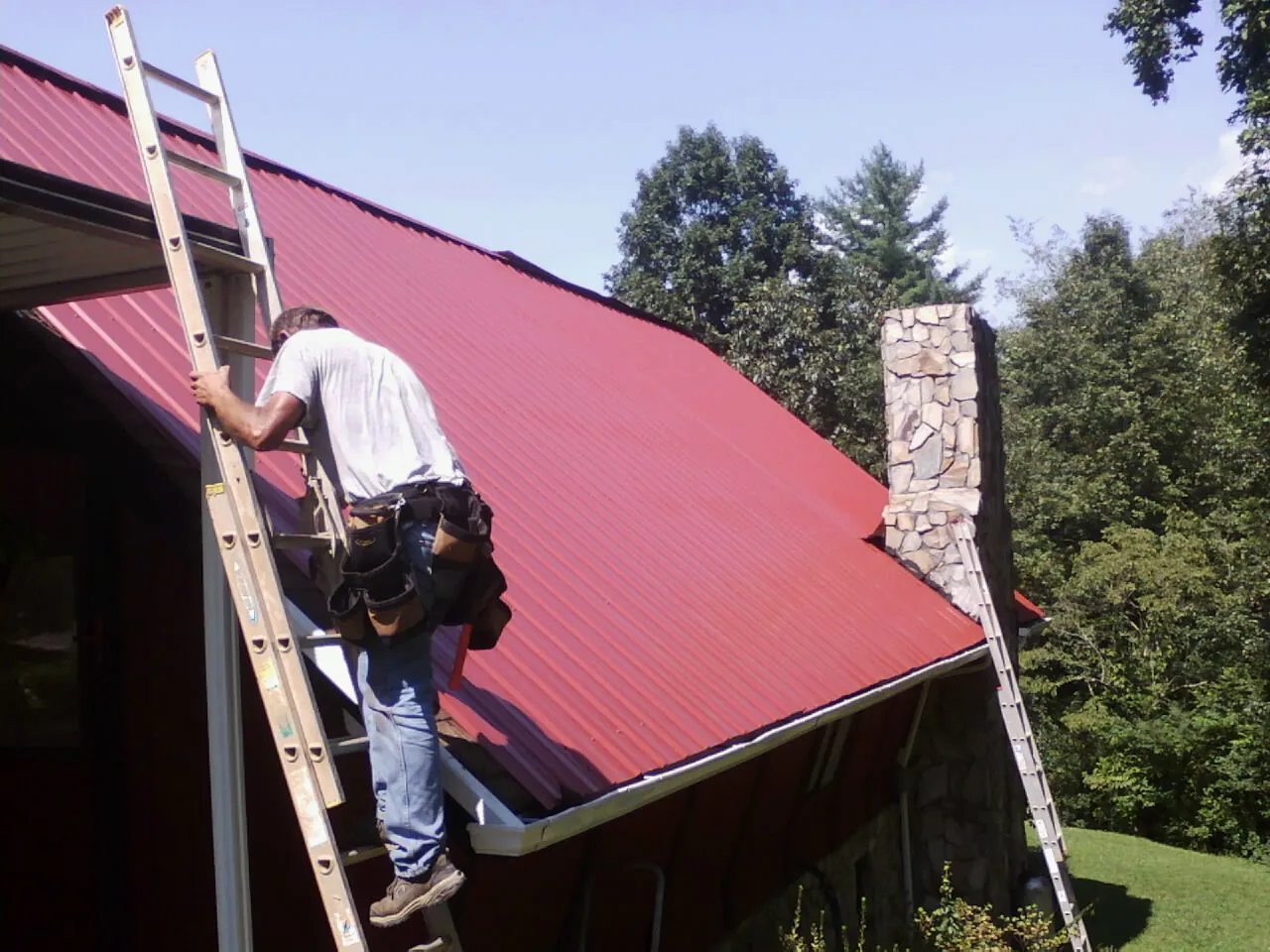 Roofer on a ladder beside a red metal roof and stone chimney — Soffit & Fascia Repair by Roofing Brothers in Bradley